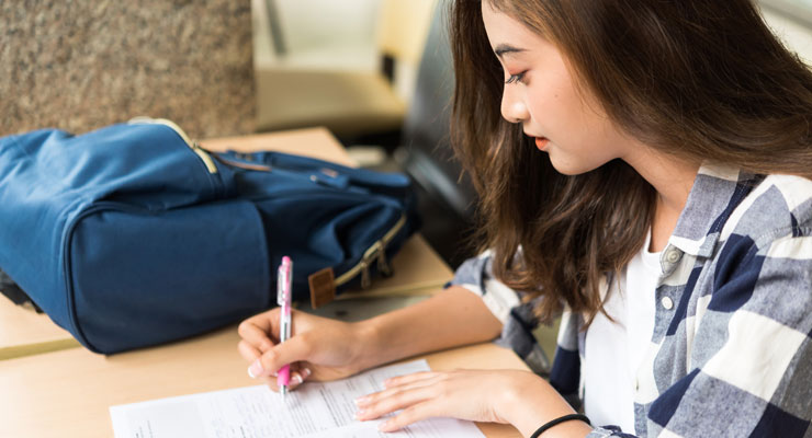 A student writing on a printed form at a wooden desk, a navy backpack beside her